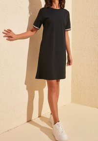 Černé šaty s krátkými rukávy zdobené červenobílými pruhovanými detaily. Jednoduchý design, délka ke kolenům, nošeno s bílými teniskami na texturovaném pozadí.