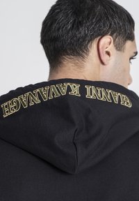 Sudadera negra con cordón y texto bordado en dorado en el cuello que dice "GA NNI KAVANAGH". Textura suave y ajuste casual.