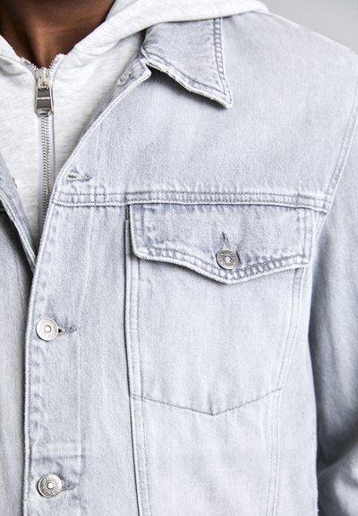 Lichtgrijze denimjack met een gestructureerd ontwerp, voorzien van een puntige kraag, een borstzak aan de voorkant en zilveren hardwareaccenten.