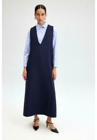 Robe sans manches bleu marine avec un décolleté en V profond, associée à une chemise boutonnée bleu clair en dessous. Présente une silhouette fluide et des talons aiguilles noirs pointus.