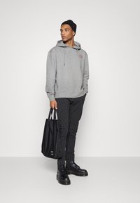 Grauer Hoodie mit Kordelzug, gestreifte schwarze Hose, schwarze Stiefel und eine schwarze Tragetasche. Der Hoodie hat ein kleines Logo.