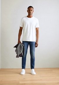 White short-sleeve t-shirt, blue jeans, and white sneakers. Gray jacket draped over left arm; standing on wooden floor against a light wall.