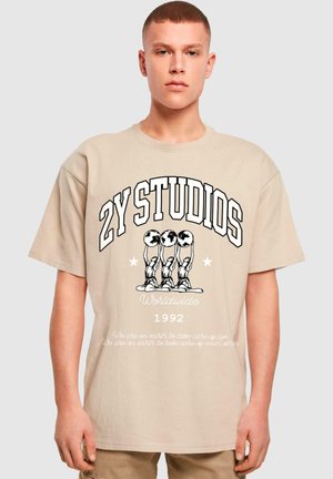 Beige oversized bomull t-skjorte med "2Y STUDIOS" i fet svart tekst, grafikk av tre hender som holder globuser, og hvit tekst under.