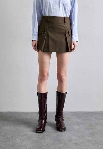 Mini jupe marron avec poches latérales, associée à des bottes en cuir laquées bordeaux jusqu'aux genoux, ornées d'un embossage floral et d'accents en métal argenté.