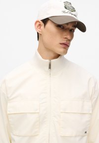 Giacca beige chiaro con collo alto, due tasche frontali e chiusura con cerniera. Cappellino bianco con logo ricamato e visiera marrone.