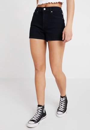 Mujer con pantalones cortos de mezclilla negros, top corto blanco y zapatillas negras de caña alta con cordones blancos, de pie sobre un fondo blanco.