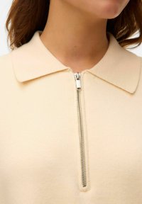 Beige polo-tröja med dragkedja och mjuk textur, med en traditionell krage och en silverfärgad dragkedja vid halslinningen.