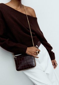 Rjava pletena jopica z izrezom na ramenih, nošena čez bele hlače, kombinirana s temno rjavo torbico z zlatimi poudarki in teksturo čez ramo.
