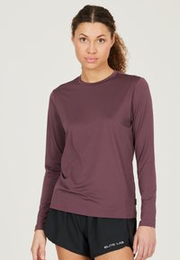 Langärmliges, lila Sportshirt aus einem glatten, leichten Stoff mit Rundhalsausschnitt und dezenten Nähten. Kombiniert mit schwarzen Shorts.