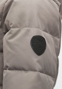 Pufová bunda v tlmených šedých farbách s prešívanou textúrou. Súčasťou je čierna nášivka v tvare štítu s názvom značky "ONLY."