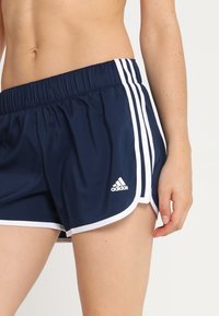 Kvinna i marinblå Adidas-träningsshorts med vita räfflor och logotyp, står med vänster arm avslappnad vid sin sida.