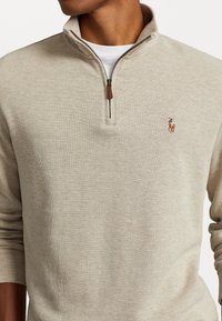 Beige stickad tröja med 1/4 dragkedja, utrustad med en läderdragflik och en broderad logga på vänster bröst. Mjuk textur och avslappnad design.