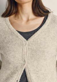 Hele beež kootud kardigan tekstuurse viimistlusega, V-kaelus, kaks nuppu, kantud tumehallal topil. Pehme, sodi materjal.