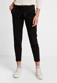 Pantalon noir avec taille à cordon de serrage et détails à pompons, coupe ajustée, longueur jusqu'à la cheville, confectionné en tissu de coton doux. Associé à des baskets blanches.