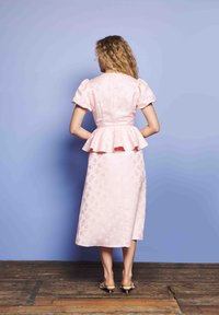 Γυναίκα με σγουρά μαλλιά στέκεται μπροστά σε έναν λευκό τοίχο, φορώντας ένα ροζ υφασμάτινο φόρεμα με φουσκωτά μανίκια και μεσαία ζώνη.
