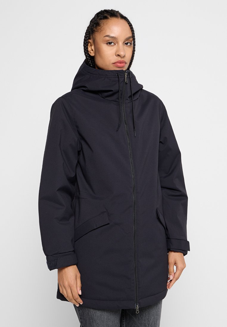 Frau mit geflochtenem Haar, die einen schwarzen Wintermantel mit Reißverschluss und Kapuze trägt, ausgestattet mit Klappentaschen und verstellbaren Bündchen, steht vor einem schlichten Hintergrund.