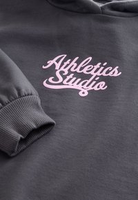 Tummanharmaa huppari, jossa on resorihihansuut ja rintakehässä vaaleanpunainen, käsinkirjoitustyyppinen teksti "Athletics Studio".