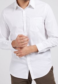 Witte blouse met lange mouwen en knopen, met een gebogen zoom, een enkele borstzak en een gladde katoenstructuur. De mouwen zijn voorzien van knoopmanchetten.