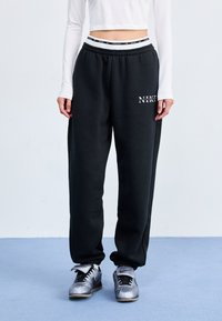 Pantalones de chándal negros con cintura elástica, que presentan un logo blanco de Nike. Combinados con zapatillas deportivas plateadas, de pie sobre una superficie azul.