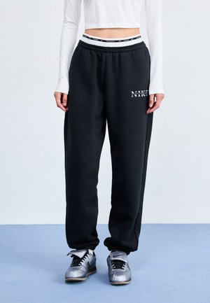 Black sweatpants with an elastic waistband, featuring a white Nike logo. Paired with silver athletic shoes, standing on a blue surface.