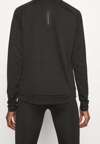 Musta pitkähihainen urheilupaita, jossa on kiinteä muotoilu, sileä pinta ja hienovaraiset heijastavat yksityiskohdat selän keskiosassa.