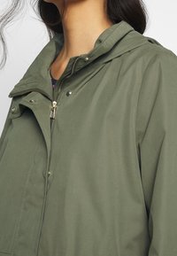 Kapuce, olivově zelená bunda má zipový přední díl, patentky a měkký, strukturovaný povrch s viditelnými švy a kapsami.