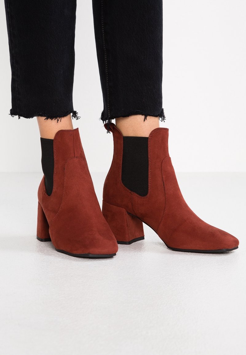 Bottes chevilles en suede marron avec talons carrés portées avec un pantalon noir à ourlet effiloché sur un fond de sol blanc.