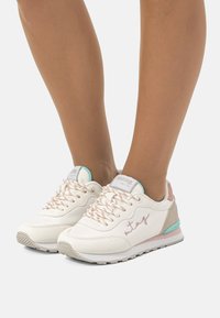 Zapatillas deportivas blancas con detalles en rosa, cuero texturizado, ribete azul pastel y cordones con estampado, que cuentan con un logo discreto en el costado.