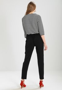 Woman standing facing away, wearing black pants, black-and-white striped long sleeve top, and red high-heeled shoes.