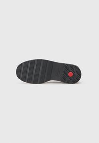 Semelle de chaussure en caoutchouc noir avec des rainures horizontales et verticales, présentant un accent circulaire rouge près du talon et des motifs de grip texturés.