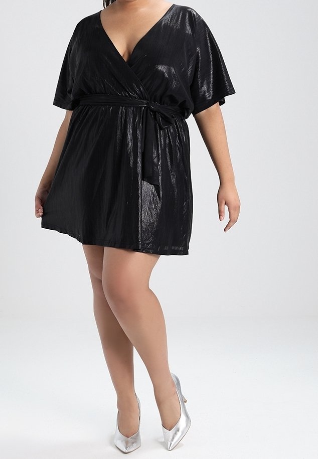 Femme portant une robe portefeuille noire brillante à manches courtes et des chaussures à talons hauts argentées, debout devant un fond clair uni.