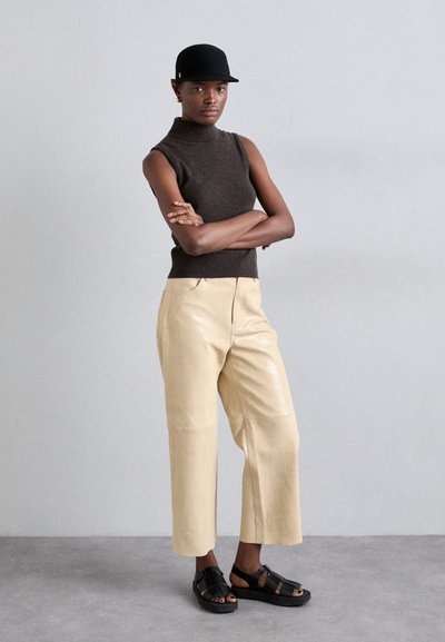 Top sin mangas de color marrón con cuello tipo mock, pantalones de pierna ancha brillantes en color beige, gorra negra y sandalias negras con tiras. La modelo está de pie con los brazos cruzados.