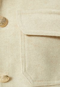 Beige uldblanding jakke detalje med en guldbutton og en brystlomme med en ren, tekstureret overflade.