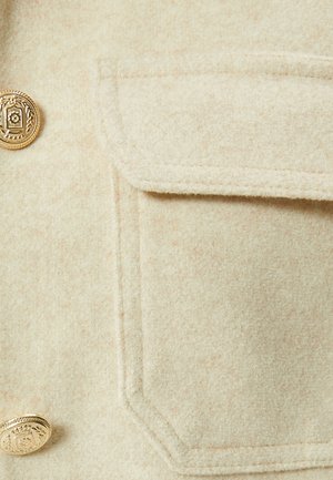 Beige wool-blend jacket detail featuring a gold button and a chest pocket with a clean, textured surface.