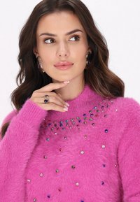 Fuchsia sweater med tekstureret, fluffy materiale; pyntet med multicolore rhinsten. Model accessorize med statement øreringe og ring.