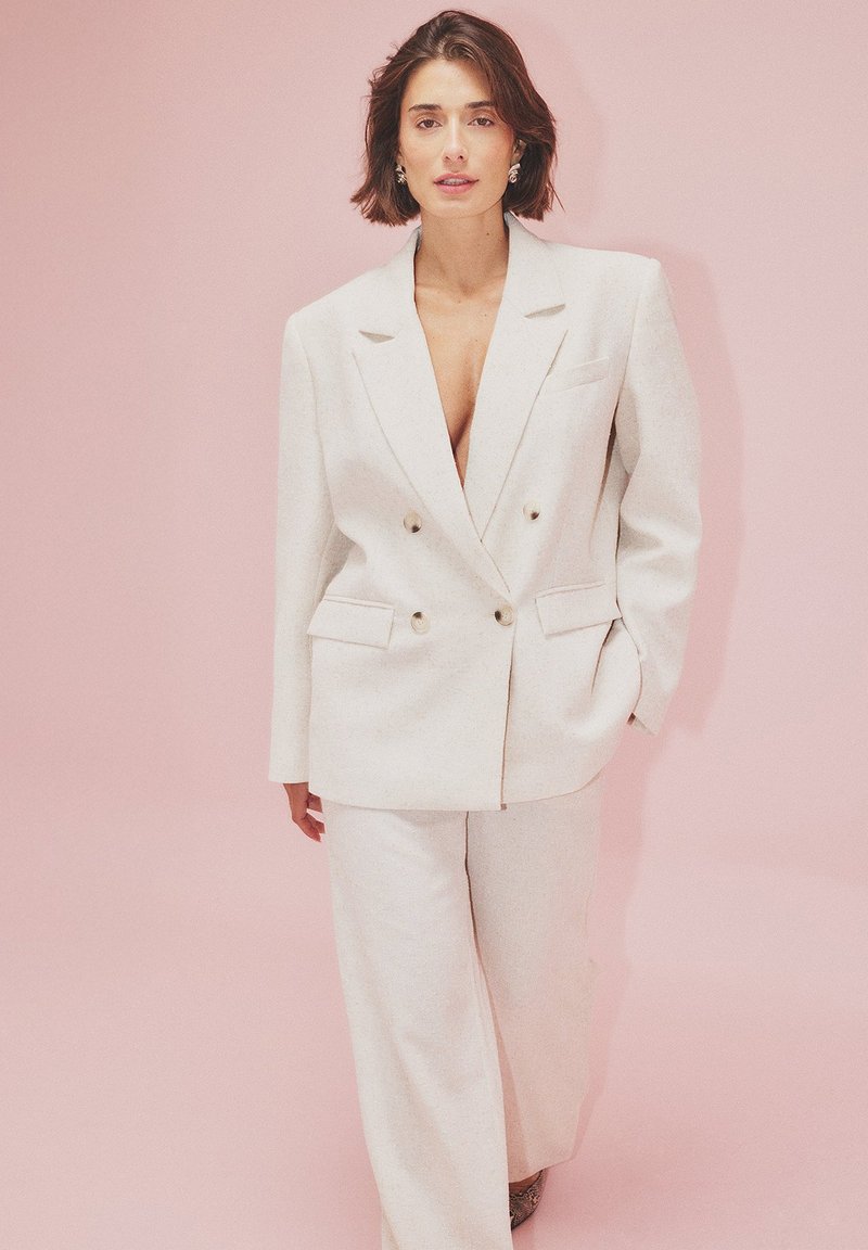 Blazer double boutonné de couleur claire avec deux poches avant, associé à un pantalon à jambes larges. Texture lisse, avec un col à cran et de grands boutons.