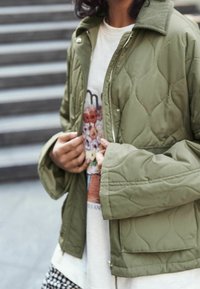 Person in einer olivgrünen Steppjacke über einem weißen T-Shirt mit floralem Grafikdruck, der die Jacke in der Nähe einer Treppe anpasst.