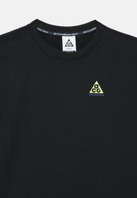 Μαύρο βαμβακερό μπλουζάκι με στρογγυλή λαιμόκοψη, που διαθέτει μικρό κίτρινο τρίγωνο λογότυπο με το κείμενο «ACG» και «All Conditions Gear» σε μπλε χρώμα.