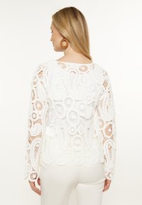 Bílý háčkovaný top s dlouhými rukávy, který obsahuje propracované květinové a paisley vzory. Zadní design je průsvitný s vlnitým lemem.