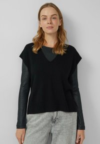 Czarny dziergany sweter bez rękawów z dekoltem w serek, z prążkowanym dołem i teksturowaną warstwą pod spodem. Noszony na ciemnej, błyszczącej bluzce z długim rękawem.