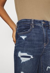 Mörkblå denimjeans med slitna områden och trasiga lappar. Har en medelhög midja och klassisk femficksdesign.
