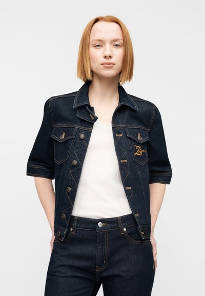 Σκούρο τζιν μπουφάν με κοντά μανίκια, διακοσμημένο με κουμπιά σε χάλκινη απόχρωση και τσέπες στο στήθος. Φοράται πάνω από ένα λευκό τοπ και συνδυάζεται με σκούρο τζιν παντελόνι.