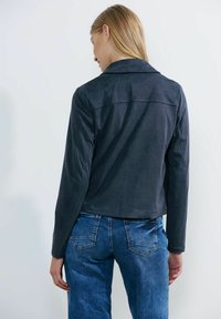Giacca in suede blu con colletto e maniche lunghe, taglio corto, texture liscia e dettaglio cucito sul retro, abbinata a jeans blu.