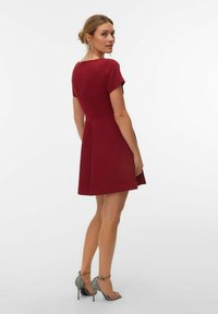 Crvena haljina kratkih rukava s pripijenim gornjim dijelom i proširenim suknjom, s okruglim izrezom i patentnim zatvaračem na leđima. Nošena sa srebrnim sandalama na visoku petu.