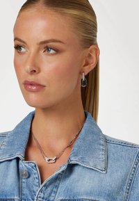 Collana in argento con pendente geometrico, abbinata a orecchini pendenti coordinati. La modella indossa una giacca di jeans chiara.