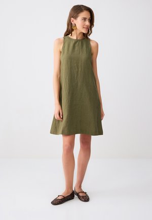 Młoda kobieta stoi, ubrana w oliwkową, bezrękawnikową sukienkę do kolan, wyraziste kolczyki oraz ciemne, plecione buty płaskie, spogląda w prawo.