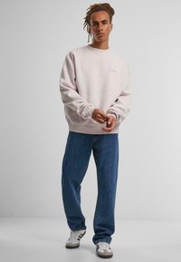 Lys rosa sweatshirt med rund hals og logodetalj, kombinert med løstsittende blå jeans og hvite joggesko. Den myke stoffoverflaten er synlig.