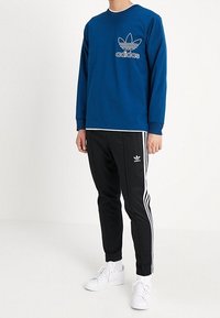 Niebieska bluza z długim rękawem z białym logo Adidas na piersi, zestawiona z czarnymi zwężanymi dresami z białymi paskami po bokach i logo.