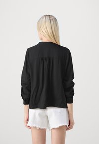 Schwarze Bluse mit langen Ärmeln und gerafft am Rücken, aus glattem Stoff. Kombiniert mit weißen Shorts mit ausgefranstem Saum.
