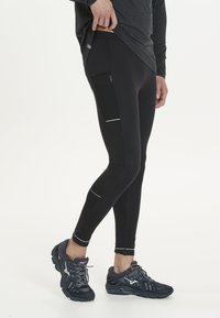 Sorte atletiske leggings med en tætsiddende pasform, der har en mesh-lomme på siden og reflekterende detaljer, parret med mørke løbesko.